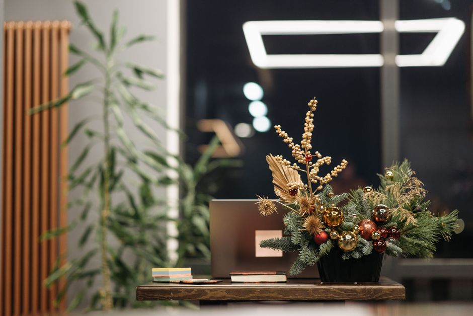 A festive floral arrangement featuring a mix of evergreen branches, gold and red baubles, dried thistles, and small berries, elegantly displayed in a black vase on a wooden table. The display includes