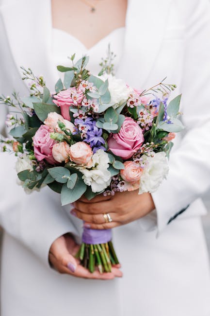 A person dressed in a white outfit holds a vibrant bridal bouquet featuring large pink roses, pale peach spray roses, and white lisianthus, accented with soft green eucalyptus leaves and purple delphi