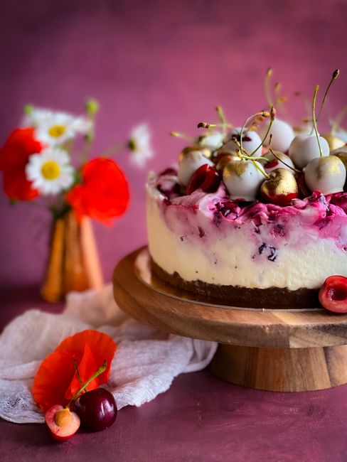 A close-up of a layered cheesecake with a pink swirl topping, decorated with white and gold cherries, placed on a wooden cake stand. In the background, a bouquet of fresh flowers in a small vase inclu