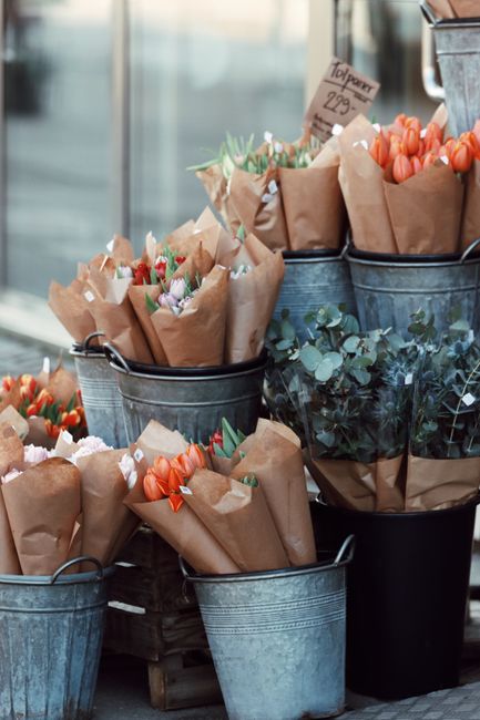 An outdoor display of fresh flower bouquets arranged in metal buckets and containers, featuring a variety of flowers including vibrant orange tulips, delicate pink roses, and small purple blossoms, al